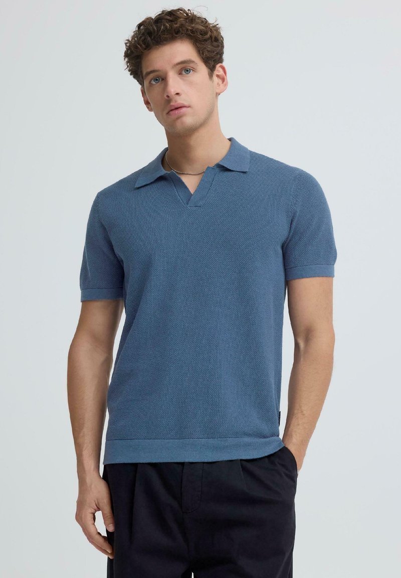 Νεαρός άνδρας με σγουρά μαλλιά, ντυμένος με μπλε πλεκτό πόλο μπλουζάκι με κοντά μανίκια και σκούρο παντελόνι, στέκεται με το ένα χέρι στην τσέπη.