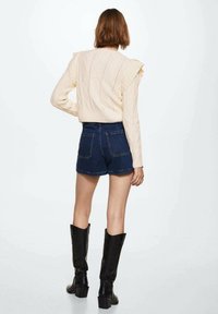 Cremiger, kabelgenähter Pullover mit rüschigen Schultern, kombiniert mit hochgeschnittenen, dunklen Denim-Shorts und schwarzen, knielangen Stiefeln.