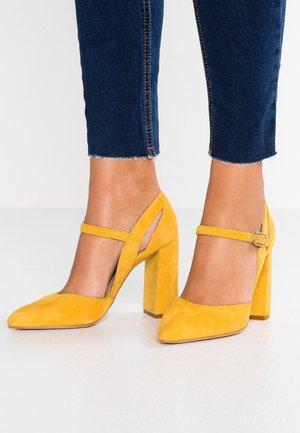 Žluté nubukové lodičky na vysokém podpatku se špičatou špičkou, řemínkem kolem kotníku a geometrickým vykrojením. Robustní blokový podpatek a texturovaný povrch.
