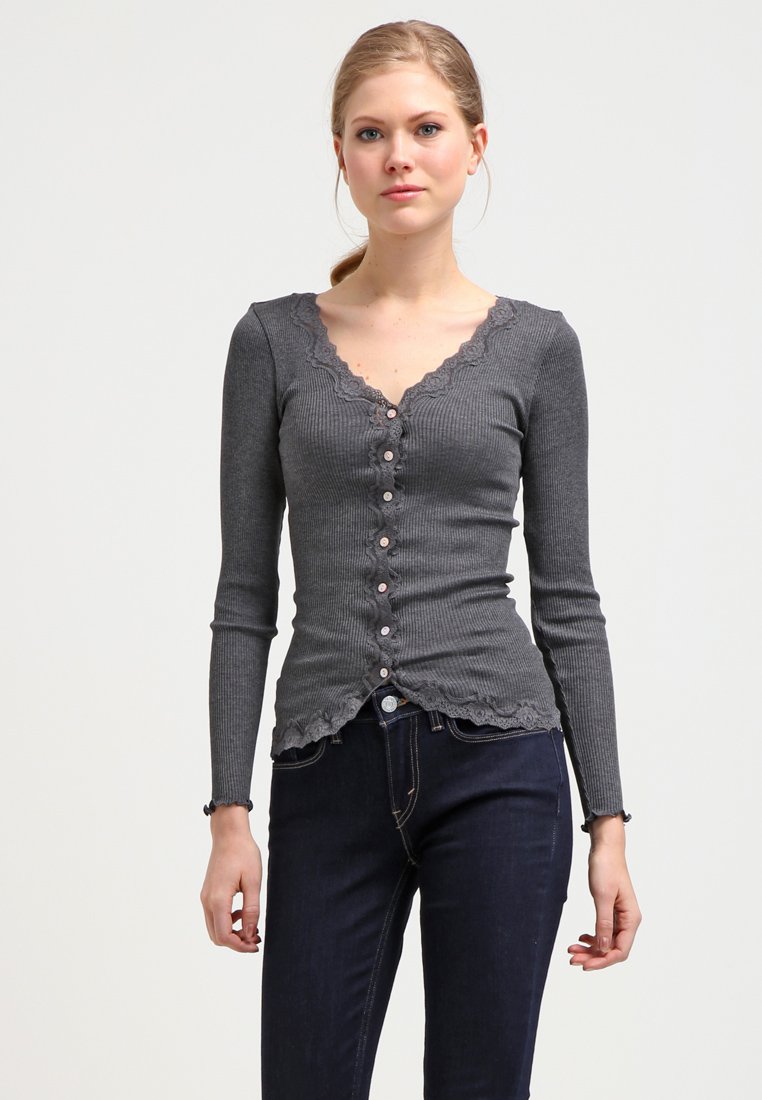 Grijze geribbelde cardigan met lange mouwen, een V-hals, golvende randen en roze knoopsluitingen. Gecombineerd met donkere spijkerbroeken.