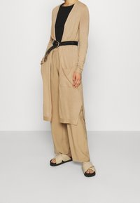 Beżowy kardigan na czarnym topie, przewiązany czarnym paskiem. Luźne beżowe spodnie i jasnobeżowe sandały z czarną podeszwą.
