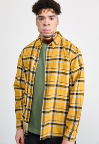 Camisa de franela a cuadros amarillos y negros sobre una camiseta de cuello redondo verde. La camisa cuenta con botones y un bolsillo en el pecho. Textura suave.