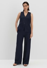 Marineblauwe mouwloze blazer met drie knopen, gecombineerd met hoge, wijde broekspijpen. Glad stof met een subtiele glans.