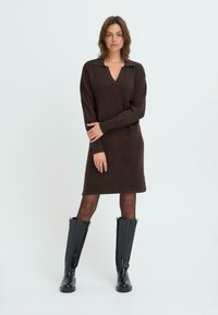 Brown knitted dress with a V-neck and collared design. Long sleeves and knee-length, paired with tall black boots. Smooth texture, minimal details.