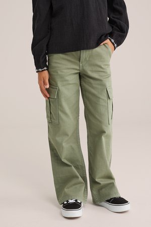 Pantalon cargo vert avec une coupe décontractée, doté de poches latérales et d'une texture en coton lisse, associé à des baskets noires et un haut sombre.