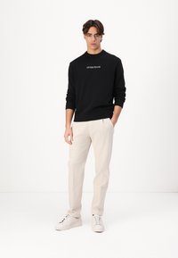 Fekete kötött pulóver "GUESS JEANS" logóval, világos bézs szabott nadrággal és fehér sportcipővel egy semleges háttér előtt.