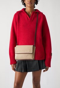 Červený pletený sveter s golierom, skombinovaný s béžovou textúrovanou crossbody taškou so zapínaním na klapku a hnedým lemovaním. Čierna plisovaná sukňa.