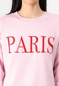 Svetloružová mikina s výrazným červeným vyšitým textom "PARÍŽ" cez hruď. Pružný výstrih, mäkká textúra a voľný strih.