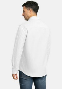 Camisa blanca de manga larga con cuello abotonado, costuras visibles y dobladillo redondeado. Combinada con jeans azules.