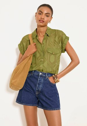 Žena v olivovo zelenej vzorovanej krátkoročnej košeli a modrých rifľových kraťasoch drží béžovú semišovú tašku s náramkami na zápästí.