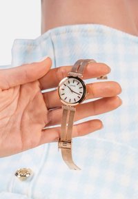 Zlata in srebrna mrežasta ura z okroglo belo površino, črnimi označevalniki ur ter logotipom 'GUESS'. Nastavljiv sponka za varno prileganje.