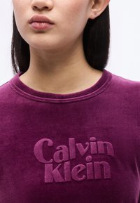 Κοντινό πλάνο ενός ατόμου που φοράει μωβ βελούδινο τοπ με ανάγλυφο κείμενο «Calvin Klein» στο στήθος, πάνω σε λευκό φόντο.