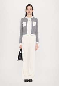 Harmaa neulecardigan, jossa on valkoisia yksityiskohtia ja taskuja, yhdistetty valkoisiin leveälahkeisiin housuihin ja mustiin loafer-kenkiin, pitäen pientä mustaa käsilaukkua.