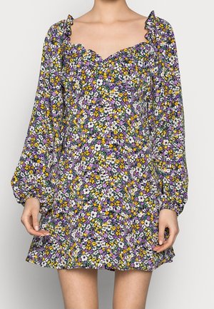 Vrouw die een lange mouwen, bloemige minijurk draagt met paarse, gele en witte bloemen en een gerimpelde vierkante halslijn.