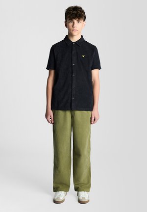 Adolescent garçon debout face à nous, portant une chemise noire à manches courtes avec boutons, un pantalon vert olive et des baskets blanches.
