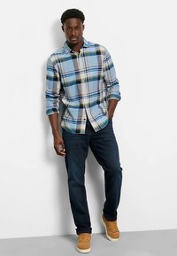 Kariertes Hemd in Blau, Weiß, Grün und Rot mit Hemdkragen zum Knöpfen. Dunkle Jeans und beige Turnschuhe komplettieren das Outfit.