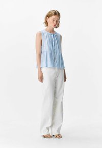 Una joven se encuentra de pie contra un fondo plano, vistiendo una blusa sin mangas de color azul claro y pantalones de pierna ancha blancos, con una expresión neutral.