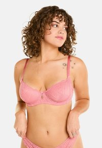 Soutien-gorge en dentelle rose avec un motif géométrique, bretelles ajustables et un petit détail décoratif au centre devant. Texture lisse.