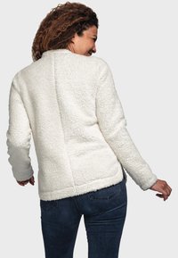 Veste en fausse fourrure couleur blanc cassé avec une finition texturée, dotée de manches longues et d'un design simple et épuré. La vue de dos montre des détails de couture.