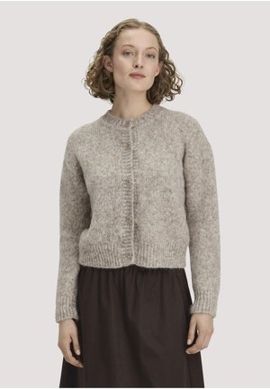 Junge Frau mit kurzen lockigen Haaren, die eine hellbraune, geknöpfte Strickjacke und einen dunklen Rock trägt, steht vor einem schlichten Hintergrund.