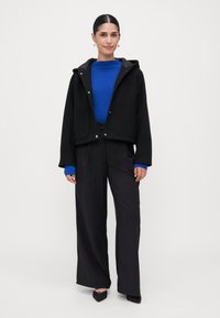 Vrouw in een zwarte hoodie, felblauwe geribbelde trui, wijde zwarte broek en zwarte puntige hakken, staand tegen een effen achtergrond.