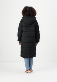 Black padded long coat with a hood, featuring horizontal stitching and a straight cut. Worn with light blue pants and beige heels.