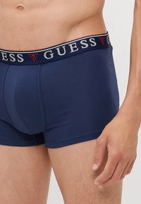 Boxeri din bumbac de culoare bleumarin cu un elastic de talie cu logo-ul GUESS în gri și roșu. Prezintă o ajustare strânsă și o textură netedă.