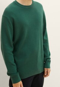 Zielony sweter z dzianiny waffle, okrągły dekolt, długie rękawy, ściągacze na mankietach i u dołu, textured materiał, noszony na ciemnych spodniach.