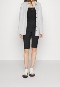 Camisa de botones de manga larga en gris claro, top negro de tirantes, pantalones cortos negros a medida y sandalias blancas de tipo slip-on con suelas negras. Texturas suaves.