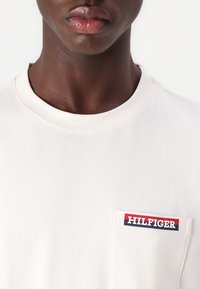 Majica kratkih rukava, s okruglim izrezom, u svijetloj krem boji s džepom na lijevoj strani prsnog dijela, na kojem se nalazi crveno, bijelo i plavo Hilfiger logo.