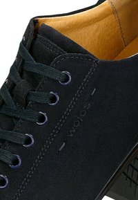 Schwarze Wildleder-Sneaker mit runden Schnürsenkeln, Metallösen und einem braunen Innenfutter. Strukturiertes Profil mit Markenlogo an der Seite.