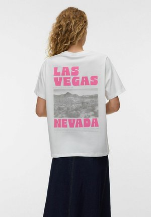 Osoba o kręconych włosach, ubrana w białą koszulkę z różowym napisem „Las Vegas Nevada” i czarno-białym zdjęciem pustynnego krajobrazu na plecach, w zestawieniu z ciemną spódnicą.