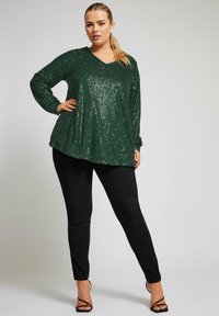 Vihreä paljettopaita pitkillä hihoilla ja V-päällä, yhdistettynä tiukkoihin mustiin housuihin ja remmisandaaleihin. Paidan pinnassa on kiiltävä, tekstuurinen pinta.