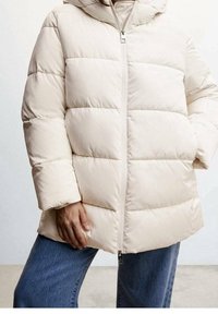 Prošívaná bunda světle béžové barvy s kapucí, s vodorovným šitím, předním zipem a volným střihem. Kombinována s džínami.