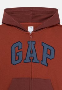 Haut de sweat à capuche zippé de couleur rouille avec de grandes lettres bleues "GAP" sur la poitrine et une étiquette de marque imprimée numériquement à l'intérieur du col.