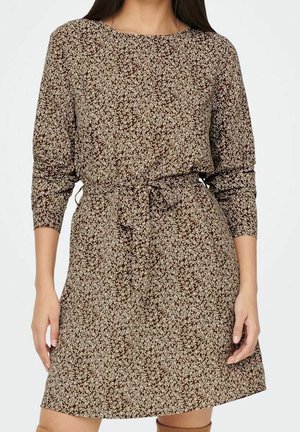 Kvinde iført en brun, langærmet blomstret kjole med et bundet taljebælte, stående med armene afslappet ned langs siderne.