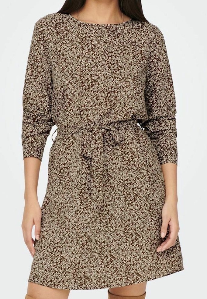 Femme portant une robe longue à manches longues marron à motifs floraux, avec une ceinture nouée à la taille, debout les bras détendus sur les côtés.