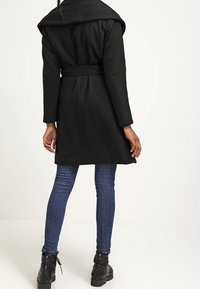 Cappotto di lana nera con ampio colletto, vita con cintura e maniche lunghe. Abbinato a jeans blu e stivaletti neri. Design semplice e elegante.