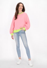 Felpa oversized rosa con polsini e orlo in verde neon, abbinata a jeans skinny blu chiaro e sneakers bianche, caratterizzata da una vestibilità rilassata.