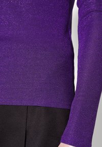 Lila ribbad långärmad topp med en strukturerad, glittrig finish, tillsammans med svart tyg nedtill. Närbild på material- och design detaljer.