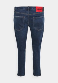 Vaqueros de mezclilla azul oscuro con un ajuste slim, bolsillos traseros y una distintiva etiqueta roja "HUGO" en la cinturilla.