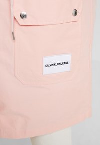 Szorty Calvin Klein Jeans w jasnoróżowym materiale, z boczną kieszenią z metalowym zatrzaskiem i białą metką z nazwą marki.