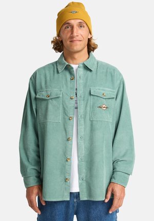 Camisa de pana color teal con dos bolsillos en el pecho, botones marrones y un parche de logo; combinada con un gorro de lana amarillo.