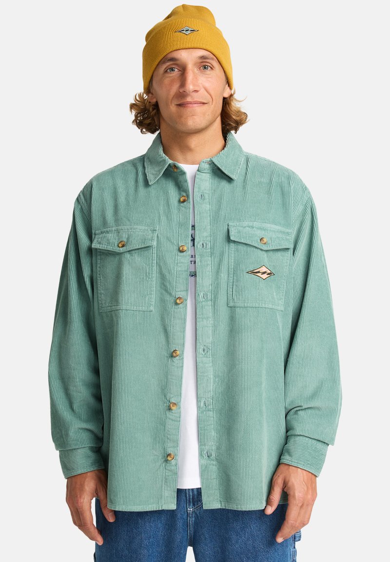 Teal corduroy shirt with two chest pockets, featuring brown buttons and a logo patch; paired with a yellow knit beanie.