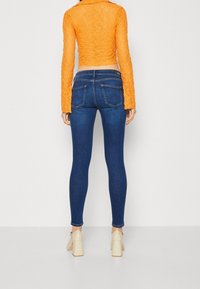 Top de encaje naranja cortado sobre unos pantalones vaqueros de mezclilla oscuros ajustados con dos bolsillos traseros. Modelo lleva zapatos de tacón de color claro.