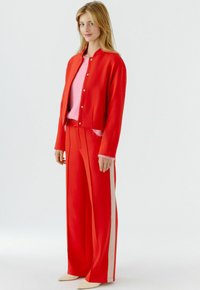 Rode jas met zilverkleurige knopen, gecombineerd met rood wijde pijpen broek met zijstrepen. Daaronder een lichtroze trui voor contrast.