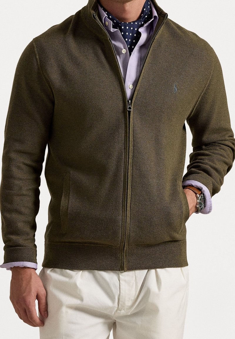 Muž v olivově zeleném zipovém svetru přes levandulovou košili s knoflíky a s kravatou s puntíky, v bílých kalhotách a s hodinkami.