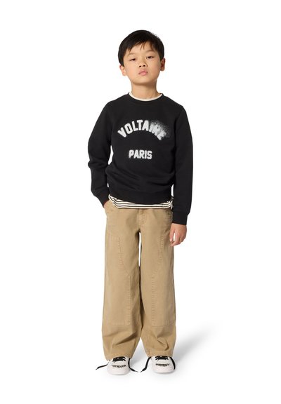 Μαύρο φούτερ με εκτύπωση "VOLTAIRE PARIS", καφέ παντελόνι με φαρδιά ίσια γραμμή και ασπρόμαυρα αθλητικά παπούτσια. Το σύνολο περιλαμβάνει πουκάμισο με ρίγες στρωμένο από κάτω.