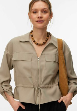 Kvinne iført beige jakke med glidelås og snor, brun perlekjede, og bærer en tan semsket skulderveske.