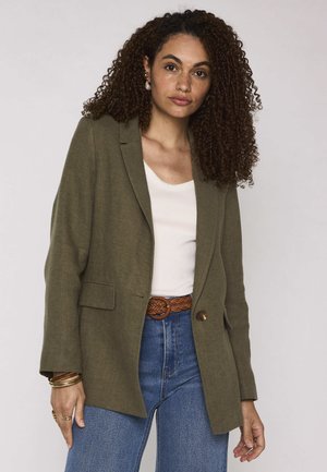 Femme aux cheveux bouclés et foncés porte un blazer vert olive, un haut blanc, un jean bleu, une ceinture tressée marron ainsi que des bracelets et bagues en or, debout devant un fond uni.